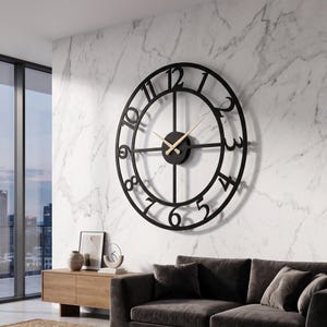 May include: A large, black, circular wall clock with a minimalist design. The clock features a black frame with cut-out numbers and a central clock face with gold-coloured hands. The clock is mounted on a white marble-patterned wall.