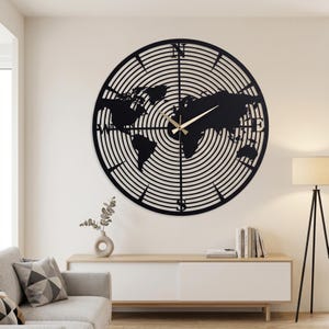 May include: A large, black metal wall clock featuring a world map design. The clock face has concentric circles and compass points. Gold-colored clock hands indicate the time. The clock is mounted on a light-colored wall.
