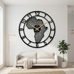 May include: A large, black metal wall clock featuring the shape of Africa with a fingerprint design. The clock hands are gold, and the numbers are cut out around the outer edge. The clock is mounted on a white wall above a white sofa.