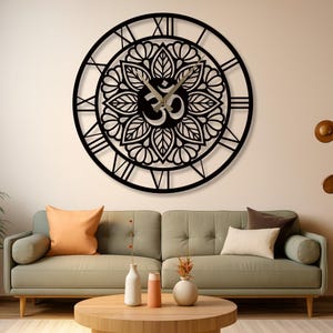 Op de afbeelding: Een grote, zwarte, ronde wandklok met Romeinse cijfers en een decoratief bloemmotief. De klok heeft het Om-symbool in het midden. De klok is gemonteerd op een lichtgekleurde muur boven een groene bank.