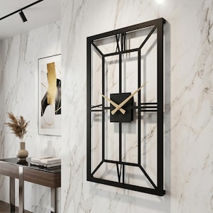 Op de afbeelding: Grote, rechthoekige wandklok met een zwart metalen frame en Romeinse cijfers. De wijzerplaat is een zwarte vierkant met goudkleurige wijzers. De klok is bevestigd aan een witte marmeren muur.