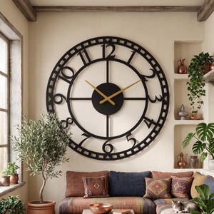 May include: A large circular wall clock featuring a black metal frame with open-work detailing and bold black Arabic numerals. The clock has a solid black center disc and two thin, gold-colored hands indicating the time against the wall.
