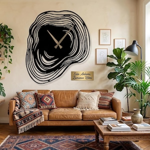 May include: A large, black, abstract wall clock with a unique, layered design. The clock hands are a light brown color. The clock is mounted on a beige wall above a brown leather sofa with decorative pillows. A small sign below the clock reads "Evin Anlamı, Zamanın Ritmi."