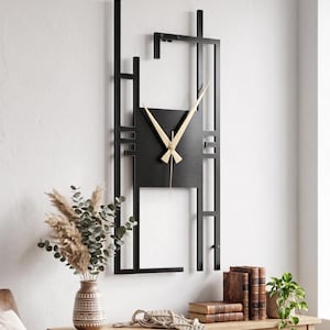 May include: A contemporary black metal wall clock with gold hands. The clock features a geometric design and is mounted on a white wall. The clock is part of a decorative display with a wooden table, books, and a vase of dried flowers.