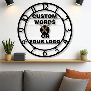 May include: Black, round wall clock with the text "CUSTOM WORDS OR YOUR LOGO". The clock has a minimalist design with black numerals and hands. The clock is mounted on a white wall.