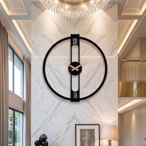 May include: A large, black, circular wall clock with Roman numerals at the top and bottom. The clock hands are a light wood color, and the clock is mounted on a white marble wall. A chandelier hangs above the clock.