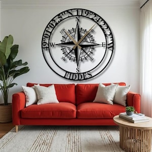 May include: A large, black metal compass rose wall clock with gold-colored hands. The clock features the text "FIDDES" at the bottom and coordinates around the outer edge. The clock hangs above a red sofa with white pillows.