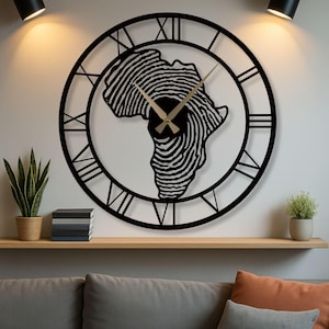 Op de afbeelding: Zwarte metalen wandklok met een vingerafdrukontwerp van Afrika. De klok heeft Romeinse cijfers en gouden wijzers. Gemonteerd op een witte muur boven een houten plank. De diameter is ongeveer 60 cm.