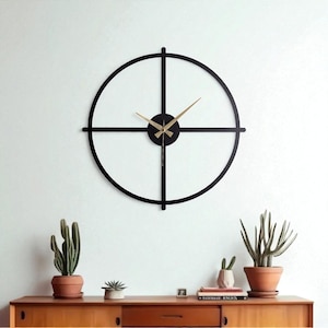 Puede incluir: Un reloj de pared moderno de metal negro con un diseño circular y manecillas doradas. El reloj presenta un diseño en forma de cruz dentro del círculo. Está montado en una pared blanca sobre un mueble de madera.