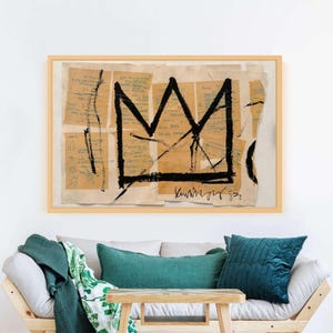 Puede incluir: Obra de arte enmarcada con un dise&ntilde;o de corona negra sobre un collage de fragmentos de papel beige con notas manuscritas. La obra est&aacute; en un marco de madera clara. Se exhibe sobre un sof&aacute; de madera clara con cojines verde azulado y una manta verde.