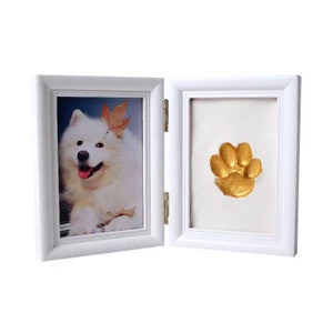 May include: White double picture frame with a photo of a white dog in one frame and a gold paw print in the other frame.