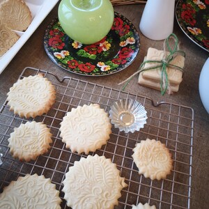 May include: A close-up of a wire rack with a variety of biscuits on it. The biscuits are round and have a decorative design on them. There are also a few biscuits on a plate in the background.