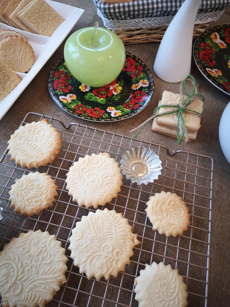 May include: A close-up of a wire rack with a variety of biscuits on it. The biscuits are round and have a decorative design on them. There are also a few biscuits on a plate in the background.
