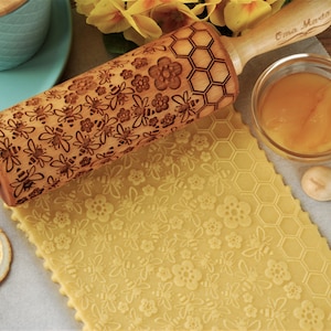 May include: A wooden rolling pin with a carved design of bees, flowers, and honeycomb. The rolling pin is on a piece of dough with a similar design. The text "Cma Marta" is on the handle of the rolling pin.