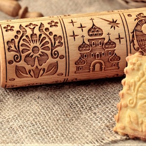 May include: A wooden rolling pin with intricate carvings of a flower, a church, and a rooster. The rolling pin is on a burlap surface next to a stack of cookies with a floral design.