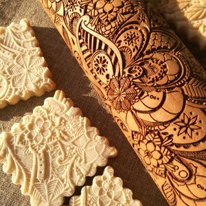 May include: A wooden rolling pin with an intricate floral design carved into the surface. The rolling pin is used to create decorative patterns on cookies and other baked goods. The image also shows several cookies with the same floral design.