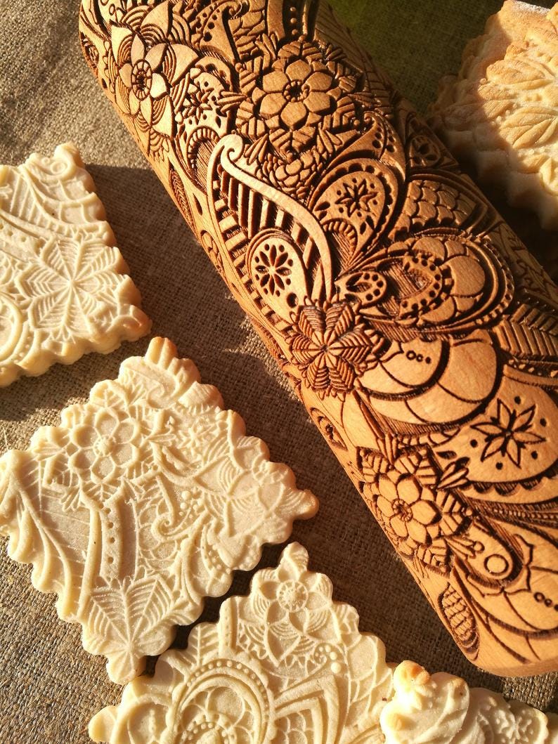 May include: A wooden rolling pin with an intricate floral design carved into the surface. The rolling pin is used to create decorative patterns on cookies and other baked goods. The image also shows several cookies with the same floral design.