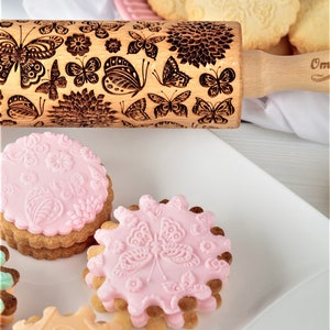 May include: A wooden rolling pin with a butterfly and floral design. The rolling pin is used to create decorative patterns on cookies. There are several cookies with pink icing and butterfly designs on a white plate.
