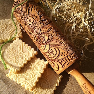 May include: A wooden rolling pin with an intricate floral design engraved into the surface. The rolling pin has a wooden handle with the words "Emma Maria" engraved on it. The rolling pin is surrounded by several cookies with the same floral design.