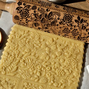 Può includere: Un mattarello di legno con un motivo floreale inciso sulla superficie. Il mattarello viene utilizzato per stendere la pasta per biscotti, che presenta un motivo floreale simile.