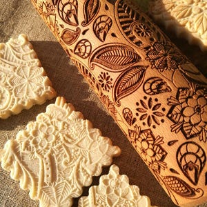 May include: A wooden rolling pin with a carved floral design. The rolling pin is used to create decorative patterns on cookies and other baked goods. The image also shows several cookies with the same floral design.
