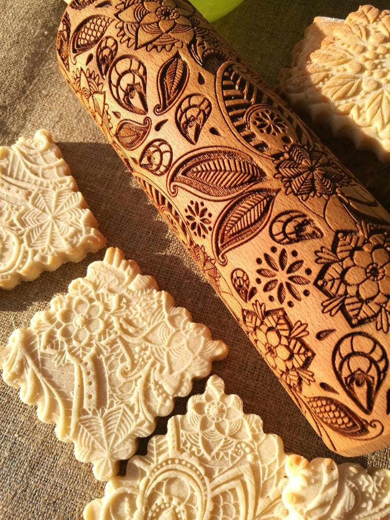 May include: A wooden rolling pin with a carved floral design. The rolling pin is used to create decorative patterns on cookies and other baked goods. The image also shows several cookies with the same floral design.