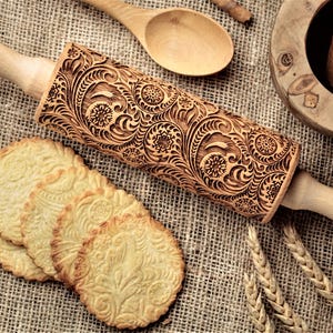 May include: A wooden rolling pin with an intricate floral design engraved on the surface. The rolling pin has a wooden handle with the text "Oma Marta" engraved on it. There are four cookies with the same floral design on a hessian surface.