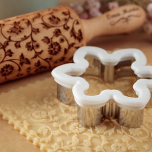 Può includere: Un mattarello di legno con un motivo floreale viene utilizzato per creare un motivo in rilievo su un foglio di pasta per biscotti. Uno stampino per biscotti in metallo a forma di fiore viene posizionato sopra la pasta.