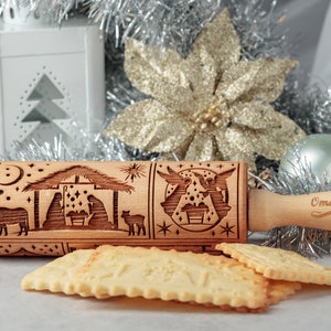May include: A wooden rolling pin with a detailed engraved nativity scene, stars, and crescent moon. The handle is engraved with the name "Oma Marta". Several Christmas cookies are in the foreground, with silver tinsel and a gold poinsettia in the background.