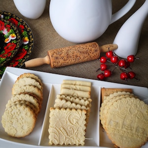 May include: A white platter with three sections holds a variety of cookies. The cookies are decorated with a floral pattern and are arranged in neat rows. A wooden rolling pin with a floral design is also visible in the image.