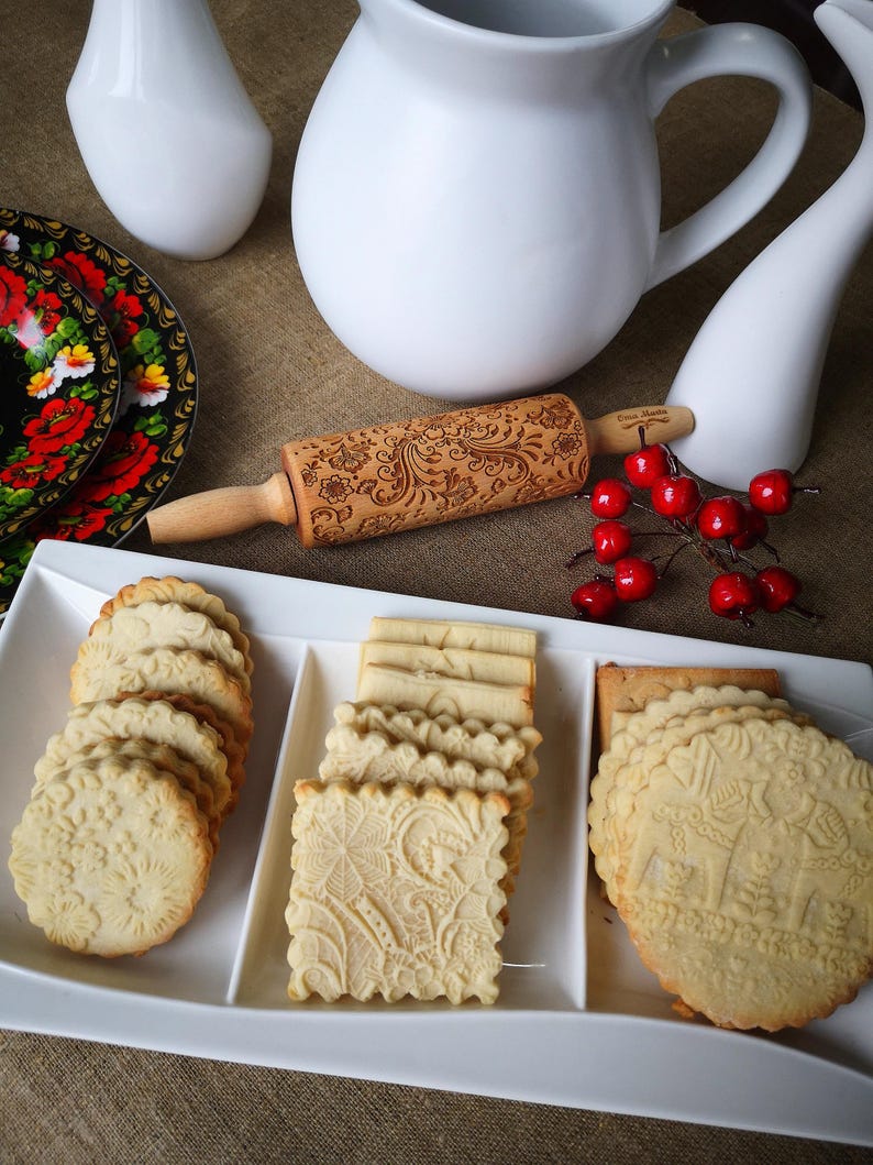 May include: A white platter with three sections holds a variety of cookies. The cookies are decorated with a floral pattern and are arranged in neat rows. A wooden rolling pin with a floral design is also visible in the image.