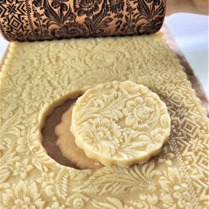 May include: A wooden rolling pin with a floral design is used to roll out dough for cookies. The dough is imprinted with the same floral design as the rolling pin. A cookie cut out in the shape of a circle is shown next to the dough.