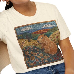 May include: Cream-colored t-shirt featuring a detailed illustration of a capybara in a landscape with flowers and rolling hills. The artwork is framed within a rectangular border. The person is wearing blue jeans.