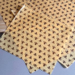 May include: A set of square beeswax food wraps featuring a repeating pattern of small brown bees on a light yellow background. The fabric sheets have pinked, zigzag edges and a smooth, slightly translucent texture characteristic of wax coating.