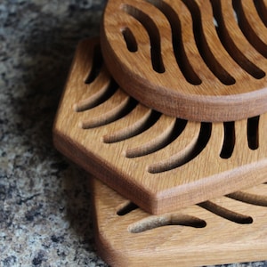 May include: Three wooden coasters with a wavy design. The coasters are stacked on top of each other. The top coaster is round, the middle coaster is hexagonal, and the bottom coaster is square.
