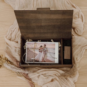 Puede incluir: Una caja de madera de color marrón oscuro con la tapa abierta que contiene una pila de fotografías rectangulares atadas con cordel. Las fotos muestran a una pareja en una playa. Una pequeña memoria USB de madera se encuentra en un compartimento.