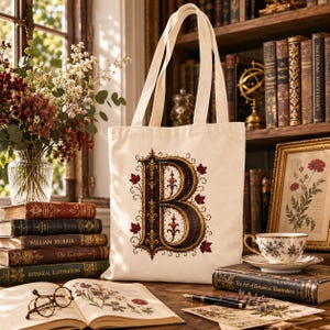 May include: A cream-colored canvas tote bag featuring a large, ornate, illuminated letter B in shades of gold, black, and deep red, decorated with intricate scrollwork and small red maple leaf accents, with two long shoulder straps.