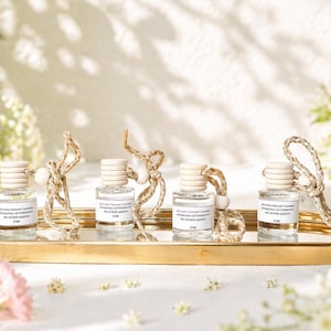 Könnte beinhalten: Vier kleine Glasflaschen mit Holzverschlüssen und Seilaufhängern liegen auf einem goldenen Tablett. Jede Flasche hat ein weißes Etikett mit schwarzem Text. Weiße Blumen und Grün umgeben die Flaschen und erzeugen eine natürliche Ästhetik.