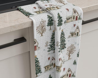 Puede incluir: Toalla de cocina blanca con un estampado de pueblo invernal. El diseño presenta ilustraciones de casas cubiertas de nieve, árboles de hoja perenne y árboles de invierno desnudos. La toalla está doblada sobre una encimera y tiene una superficie texturizada.