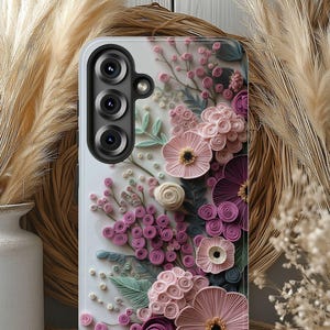 May include: A white phone case with a 3D floral design in pink, purple, and green. The case has a camera module with three lenses. The flowers are arranged on the case, creating a textured appearance.