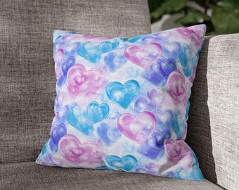 Può includere: Cuscino quadrato decorativo con un motivo di cuori traslucidi nei toni del blu, viola e rosa. I cuori sono sovrapposti su uno sfondo bianco, creando un effetto acquerello. Il cuscino è appoggiato su una poltrona grigia.