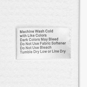 Puede incluir: Una etiqueta de tela blanca con texto negro. El texto dice: Machine Wash Cold with Like Colors, Dark Colors May Bleed, Do Not Use Fabric Softener, Do Not Use Bleach, Tumble Dry Low or Line Dry.