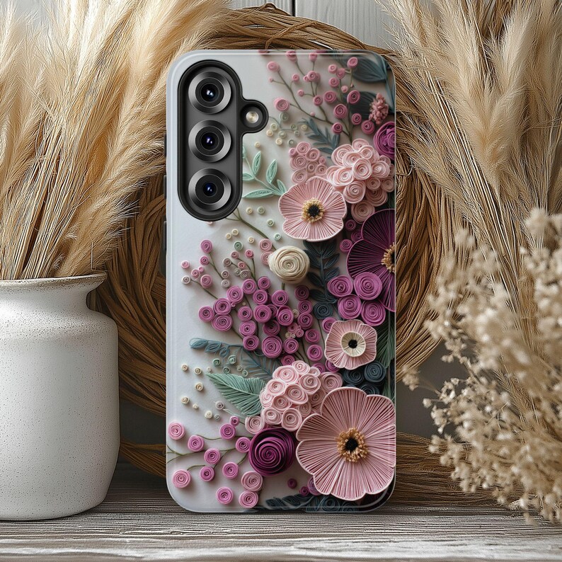 May include: A white phone case with a 3D floral design in pink, purple, and green. The case has a camera module with three lenses. The flowers are arranged on the case, creating a textured appearance.
