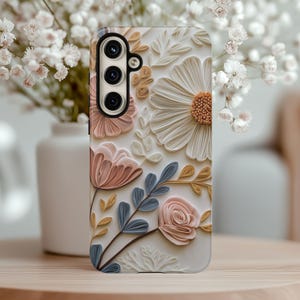 May include: A phone case with a floral design. The case features a white background with pink, blue, and brown paper flowers. The flowers are layered and have a 3D effect.