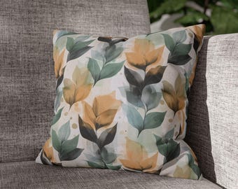 May include: A decorative square pillow with a botanical leaf pattern in shades of green, gold, and blue. The pillow is resting on a gray upholstered chair, showcasing its design and color palette. The leaves are layered on a white background.