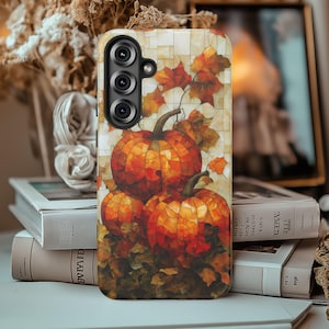May include: A phone case featuring a fall-themed design with three pumpkins in shades of orange and red, surrounded by autumn leaves. The case has a mosaic-like pattern and a camera module.