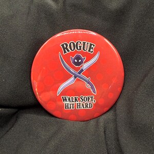 May include: A red button with the word "ROGUE" in white arched text. Below the word is a purple goblin-like character and two crossed silver swords. The text "WALK SOFT, HIT HARD" is below the swords.