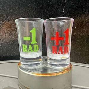 May include: Two clear shot glasses with the text "-1 RAD" in green and "+1 RAD" in red. The glasses are on a metal surface, likely for serving shots. The shot glasses are a fun novelty item.