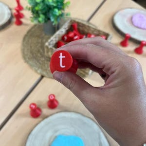 Könnte beinhalten: Ein roter, runder Stempel mit dem weißen Buchstaben „t“ wird hochgehalten. Das Wort „sat“ ist in blauer Knete eingeprägt. Weitere rote Spielfiguren und eine kleine grüne Pflanze sind auf einem Holztisch zu sehen.