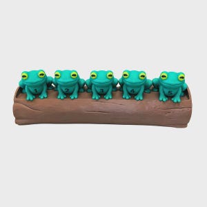 May include: Five bright green frogs with yellow eyes are positioned in a row on top of a brown, textured log. Each frog has a black number printed on its chest, ranging from one to five, displayed in sequential order from left to right.